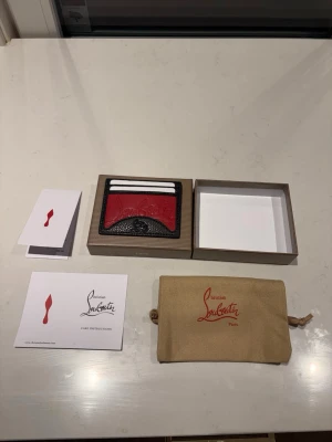  röd kortplånbok - Snygg kortplånbok i svart och rött läder. Plånboken har präglad logga på framsidan och plats för flera kort. Kommer med originalförpackning, dustbag och äkthetskort. Perfekt accessoar för dig som gillar lyxiga detaljer.