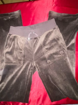 Bruna velourbyxor Juicy Couture - Mjukisbyxor i brun velour från Juicy Couture med bred resår i midjan och dragsko. Byxorna har raka ben, två framfickor och en broderad JC-logga på ena fickan. Perfekta för en chill och avslappnad stil. NYSKICK