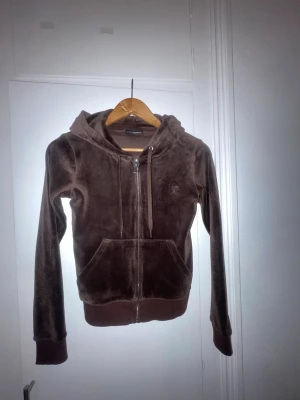 Brun velour hoodie från Juicy Couture - Mjuk och skön brun hoodie i velour från Juicy Couture. Dragkedja framtill, två fickor och justerbar huva med snören. Ikonisk JC-logga broderad på bröstet. Perfekt för en avslappnad och trendig look. NYSKICK