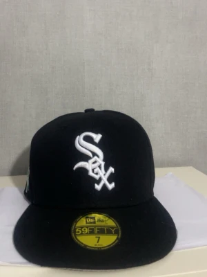 Chicago White Sox svart keps 55.8 cm - Snygg svart 59FIFTY keps från New Era med Chicago White Sox-logga broderad i vitt framtill. På sidan finns broderade detaljer med texten 'Greetings from Chicago' och gröna skyltar med 'Chicago' och 'Southside'. Klassisk flat brim och streetwear.