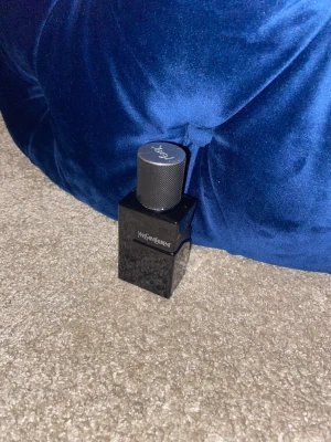 Yves Saint Laurent svart parfymflaska - Snygg parfymflaska från Yves Saint Laurent i svart glas med elegant, cylinderformad kork. Flaskan har en stilren och modern design med märkets logga på framsidan och korken. Perfekt för dig som gillar exklusiva dofter och lyxig känsla.