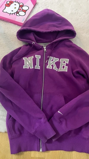 Lila hoodie med dragkedja från Nike - Snygg lila hoodie från Nike med dragkedja framtill och stora gråvita bokstäver på bröstet. Utrustad med huva och dragsko samt två fickor. Modellen är slim och passar som både XS och S