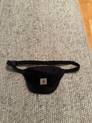 Carhartt Väska - Snygg svart axelväska från Carhartt med justerbart band och klassisk logotyp framtill. Perfekt för att bära det viktigaste och har en enkel, stilren design. Tillverkad i slitstarkt tyg och stängs med dragkedja.
