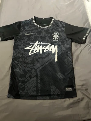 Svart Stüssy x Nike t-shirt - Säljer en svart t-shirt från Nike i samarbete med Stüssy. T-shirten har korta ärmar, vit Stüssy-logga framtill och diskret mönster i svart och grått. Rund halsringning med vita detaljer och Nike-logga på bröstet. Perfekt för dig som gillar streetwear.