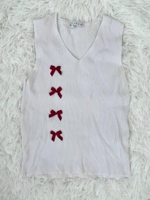 Vit ribbad topp med röda rosetter - Supersöt ärmlös topp i ribbad vit bomull med v-ringning och fyra röda rosetter på framsidan. Har små fläckar på rosetterna (se bild 3). Storlek: S, passar 34-36.