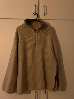 Beige stickad halvzip tröja - Stickad tröja i beige med ribbad struktur och halv dragkedja framtill. Tröjan har hög krage och vida ärmar, perfekt för lager på lager. Materialet ser ut att vara ull eller ullmix, vilket gör den extra mysig och varm.