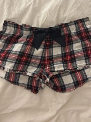 Rutiga pyjamasshorts med rosett - Söta pyjamasshorts i rött, grått och vitt rutigt mönster. Shortsen har en stor marinblå rosett i silke framtill och fransiga kanter där man ser sömmar nedtill. Tillverkade i mjukt bomullstyg, perfekta för mysiga kvällar hemma.