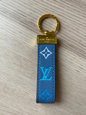  Vuitton blå nyckelring - Snygg nyckelring från Vuitton i blått läder med klassiska LV-loggor i vitt och turkost. Detaljer i guldmetall och präglad logotyp på ringen. Perfekt accessoar för att ge din nyckelknippa en lyxig touch.