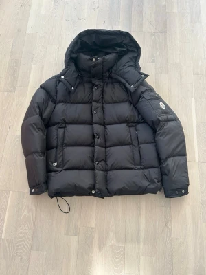 Svart dunjacka från Moncler - Snygg svart dunjacka från Moncler med avtagbar huva, stora fickor och klassisk logotyp på ärmen. Jackan har quiltad design och stängs med både dragkedja och knappar. Perfekt för kalla vinterdagar och riktigt varm tack vare dunfyllningen. MONCLER VEZERE.