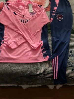 Arsenal träningsset rosa/blå Adidas - Snyggt Arsenal träningsset från Adidas i rosa och marinblått. Tröjan har halv dragkedja, långa ärmar och klubbmärke på bröstet. Byxorna har rosa detaljer och Arsenal-logga på benet. Perfekt för fotbollsträning eller chill.