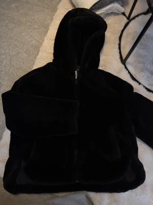 Svart teddyjacka med huva - Mysig svart teddyjacka med huva och dragkedja framtill. Jackan har en oversized passform och är gjord i fluffigt, mjukt material som ger en riktigt skön känsla. 