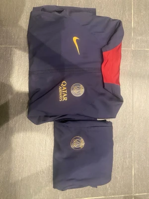 PSG träningsset från Nike, blå - Mörkblått träningsset från Nike med PSG-logga och Qatar Airways-tryck. Setet består av en jacka med gul Nike-logga och röd detalj vid axeln samt matchande byxor. Tillverkat i Dri-FIT-material som andas, perfekt för fotbollsträning.