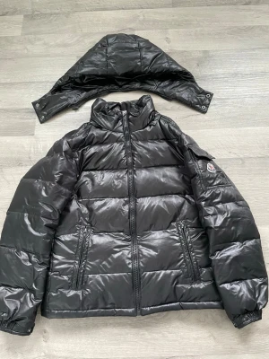 Svart dunjacka från Moncler - Snygg svart dunjacka från Moncler med avtagbar huva och två dragkedjeförsedda fickor framtill. Jackan har quiltad design, hög krage och Moncler-logga på ärmen. Perfekt för kalla vinterdagar och riktigt trendig look.