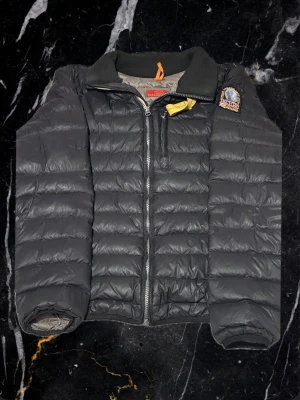 Svart dunjacka från Parajumpers söker byten - Snygg svart dunjacka från Parajumpers med klassisk quiltad design och gul detalj vid kragen. Jackan har dragkedja framtill, hög krage och patch med logga på axeln. Perfekt för kalla dagar och enkel att matcha med olika outfits. Har några defeker som trasig grej på zipen men funkar fortfarande det gula bandet saknar en grej på och den är lite sliten