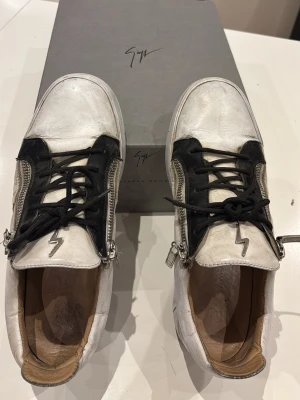 Vita sneakers från Giuseppe Zanotti - Säljer ett par vita sneakers från Giuseppe Zanotti med svarta detaljer och dragkedjor på sidorna. Skorna har en ikonisk blixtlogga på plösen, svarta skosnören och är tillverkade i läder. Snygg och modern design som sticker ut. 