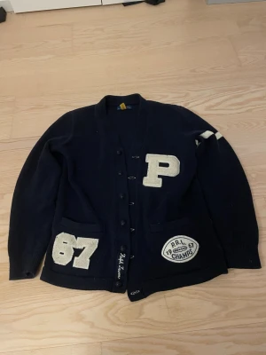 Mörkblå cardigan Ralph Lauren - Snygg mörkblå kofta i college-stil från Ralph Lauren med stora vita applikationer, bland annat bokstaven P, siffran 67 och ett märke med texten 'P.R.L. CHAMPS 1967'. Koftan har knappar framtill, två fickor och en vit rand på ena ärmen.