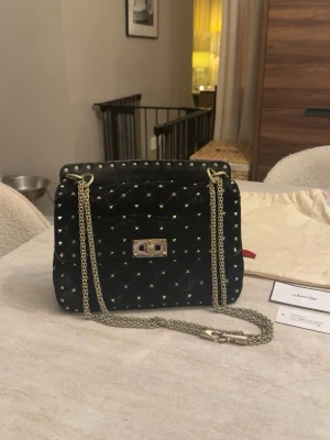 Valentino Garavani Rockstud Medium i svart sammet - Säljer min älskade Valentino Garavani Rockstud Spike i mediumstorlek, i svart sammet med guldfärgade nitar och kedja 🤍 Väskan är i jättefint skick utvändigt och sammeten är fortfarande väldigt fin. Invändigt finns det några mycket små sminkfläckar som med största sannolikhet går att få bort vid rengöring. Den kan bäras på tre olika sätt, i handen, på axeln eller crossbody. Väskan är köpt från A Retro Tale och är självklart äkta. Original dustbag samt butikskort/kvitto från A Retro Tale medföljer