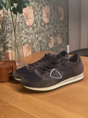 Mörkblå sneakers från Philippe Model - Snygga mörkblå sneakers från Philippe Model med detaljer i mocka och textil. Skorna har en vit sula och den ikoniska sköldloggan på sidan. Perfekta för dig som gillar stilrena och sportiga skor med en exklusiv touch.