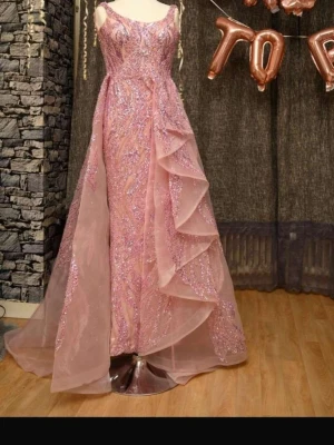 Rosa glittrig långklänning - Magisk rosa långklänning med glittriga detaljer och volanger. Klänningen har smala axelband, v-ringad urringning och lager av transparent tyg som ger extra volym. Perfekt för dig som vill sticka ut på bal eller fest.går för storlek 36 och 38