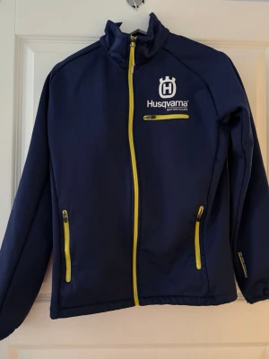 Marinblå Husqvarna jacka med gula dragkedjor - Snygg marinblå jacka från Husqvarna Motorcycles med gula dragkedjor och logga på bröstet. Jackan har hög krage, tre fickor med dragkedja och är tillverkad i ett mjukt, sportigt material. Perfekt för dig som gillar motorsport och vill ha en cool look.