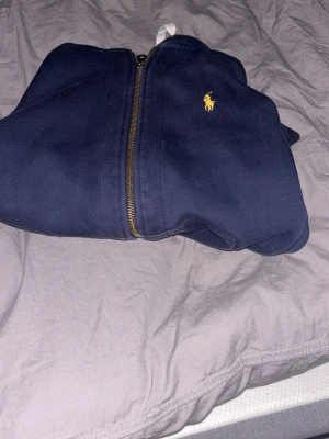 Mörkblå hoodie från Polo Ralph Lauren - Mörkblå hoodie med dragkedja från Polo Ralph Lauren. Klassisk gul logga på bröstet, huva med vita snören och ribbade muddar. Tillverkad i mjukt bomullsmaterial, perfekt för en avslappnad och stilren look. Kan sänka pris på snabb affär