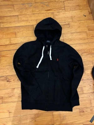 Svart hoodie från Polo Ralph Lauren - Snygg svart hoodie från Polo Ralph Lauren med dragkedja, vit dragsko i huvan och den klassiska röda loggan broderad på bröstet. Tillverkad i mjuk bomullsblandning och har två fickor framtill. Perfekt för en chill och stilren look.