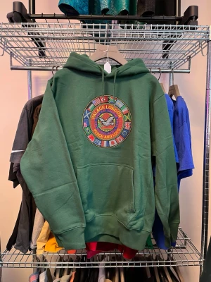Grön hoodie från Palace med brodyr - Säljer en grön hoodie från Palace med stor broderad patch på bröstet med texten 'Peace and Love' och flera flaggor runt om. Klassisk känguruficka, ribbade muddar och justerbar huva. Perfekt för dig som gillar streetwear och vill sticka ut.