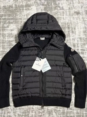 Svart dunväst med huva från Moncler - Snygg svart dunväst från Moncler med stickade ärmar och stor huva. Västen har två framfickor med knappar, dragkedja framtill och en Moncler-logga på ärmen. Materialet är en kombination av dunfyllning och stickad textil, perfekt för kyliga dagar.