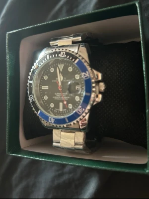 Submariner blå/silver klocka - Snygg Submariner herrklocka i silverfärgat stål med blå och svart vridring. Klockan har svart urtavla med vita markeringar och datumfönster. Klassisk länk i metall och sportig design som sticker ut.