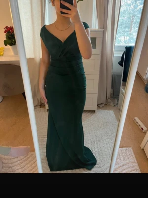 Mörkgrön långklänning med omlott - Snygg mörkgrön långklänning med omlott-detalj och v-ringning. Klänningen har korta ärmar och figurnära passform som framhäver siluetten. Perfekt för dig som vill sticka ut med en elegant och stilren look.