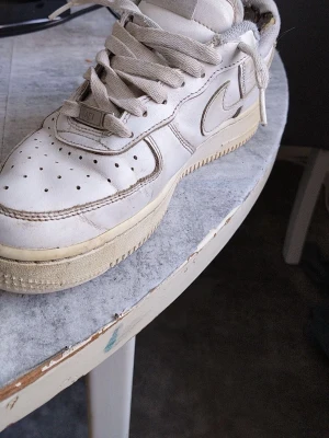 Vita Nike Air Force 1 sneakers - Klassiska vita Nike Air Force 1 sneakers i läder med perforerad tå, vit sula och ikonisk Swoosh på sidan. Skorna har låga skaft och tjocka snören. Perfekta för dig som gillar streetwear och tidlös design. Trasig på insidan och lite på sulan. Snörra om skorna själva har ej haft tid.