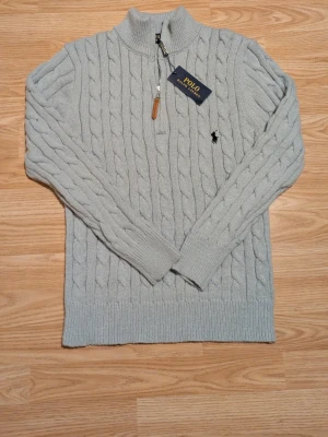 Ljusgrå kabelstickad tröja Polo Ralph Lauren - Snygg ljusgrå kabelstickad tröja från Polo Ralph Lauren med halv dragkedja och hög krage. Tröjan har ribbade muddar och nederkant samt den klassiska broderade loggan på bröstet. Perfekt för dig som gillar stilrena och tidlösa plagg.