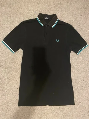 Svart piké från Fred Perry - Klassisk svart pikétröja från Fred Perry med turkosa och vita ränder på krage och ärmslut. Tröjan har korta ärmar, knappslå och den ikoniska Fred Perry-loggan broderad på bröstet. Perfekt till en avslappnad men stilren look. Otroligt snygg, några varv med en ”sån rullare” som tar bort dam är denna tröja en stark 10/10. Pris lite rörligt. Skriv om frågor!