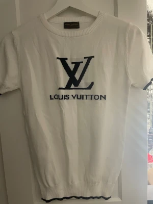 Vit Louis Vuitton t-shirt med logga - Säljer en vit t-shirt från Louis Vuitton med stor svart LV-logga och text på bröstet. T-shirten har korta ärmar, ribbad halsringning och svarta detaljer vid ärmslut och nederkant. Perfekt för dig som gillar exklusiva märken och stilren design. 💕