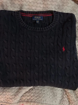 Ralph Lauren - Snygg mörkblå kabelstickad tröja från Polo Ralph Lauren med rund halsringning och röd broderad logga på bröstet. Tröjan har långa ärmar och ribbade muddar vid ärmslut och nederkant. Perfekt för dig som gillar klassisk och stilren look. Tröjan är XL men sitter som L | tveka inte på att höra av er vid minsta frågor eller fundering! :D —> Längd 58cm & bredd 46cm 