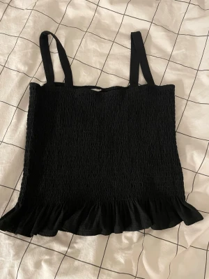 Svart smockad linnetopp med volang - Säljer en svart linnetopp med smockad struktur och breda axelband. Toppen har en söt volangkant nedtill som ger en extra touch. Perfekt att styla till jeans eller kjol för en trendig look.