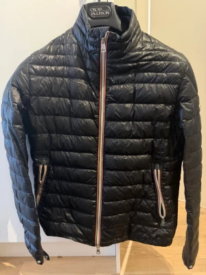 Svart dunjacka från Moncler - Snygg svart dunjacka från Moncler med quiltat mönster och hög krage. Jackan har dragkedjor med rödvita detaljer och två sidofickor. Moncler-logga på vänster ärm. Perfekt för kalla vinterdagar.
