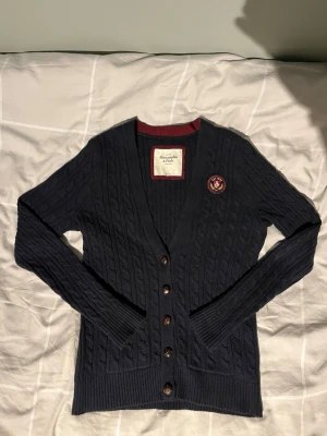 Abercrombie & Fitch Kofta - Mörkblå kabelstickad kofta från Abercrombie & Fitch med V-ringning och bruna knappar. Säljer för att den är för liten för mig🙌 Skriv om ni har några frågor! Pris kan diskuteras!