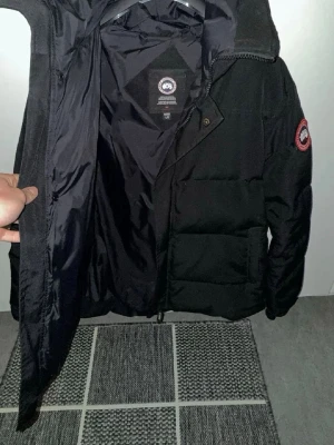 Canada Goose macmillan - Säljer en svart dunjacka från Canada Goose med klassisk patch på ärmen och dragkedja framtill. Jackan har en hög krage och quiltad design som håller värmen. Perfekt för kalla vinterdagar. Medföljer original förvaringspåse.