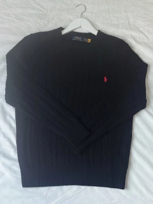 Svart kabelstickad tröja Polo Ralph Lauren - Kabelstickad svart tröja från Polo Ralph Lauren med rund halsringning och den klassiska röda logotypen broderad på bröstet. Tröjan har ribbade muddar vid ärmslut och nederkant. Perfekt för en stilren och tidlös look.