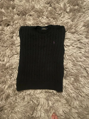 Svart kabelstickad tröja Polo Ralph Lauren - Kabelstickad svart tröja från Polo Ralph Lauren med rund halsringning och diskret broderad logga på bröstet. Klassisk modell med ribbade muddar vid ärmslut och nederkant. Perfekt för dig som gillar stilrena och tidlösa plagg.