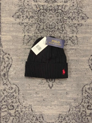 Svart mössa från Polo Ralph Lauren - Snygg svart mössa från Polo Ralph Lauren med ribbad kant och klassisk kabelstickning. Mössan har en broderad röd logga framtill och är tillverkad i mjukt material som håller dig varm. Perfekt accessoar för kalla dagar.
