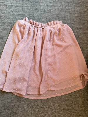 Ljusrosa kortkjol med volang - Supersöt ljusrosa kortkjol med volangkant och diskret prickigt mönster. Kjolen har resår i midjan för skön passform och är gjord i ett tunt, luftigt material som ger en flowy känsla.