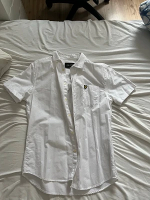 Vit kortärmad skjorta Lyle & Scott - Säljer en fräsch vit kortärmad skjorta från Lyle & Scott. Skjortan har klassisk krage, bröstficka med den ikoniska gula loggan och knappar framtill. Perfekt till sommaren och enkel att matcha med allt. Materialet är mjuk bomull.