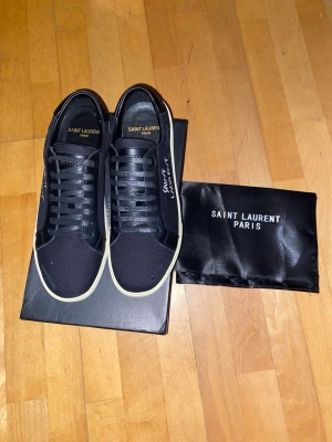 Svarta Saint Laurent sneakers - Svarta sneakers från Saint Laurent med vit sula och handskriven logga på sidan. Skorna har klassisk låg profil, svarta skosnören och detaljer i läder och canvas. Kommer med originalpåse och låda. Perfekta för en clean och stilren look.