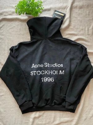 Svart hoodie Acne Studios Stockholm - Tjena! Jag Säljer en svart hoodie från Acne Studios med trycket 'Acne Studios STOCKHOLM 1996' i vitt på ryggen. Hoodien har en stor huva och ribbade muddar vid ärmslut och nederkant. Perfekt för dig som gillar streetwear och vill ha något stilrent med svensk design.