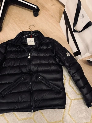 Svart Moncler pufferjacka - Säljer en svart pufferjacka från Moncler med klassisk quiltad design och två dragkedjeförsedda fickor framtill. Jackan har hög krage, lång ärm och ikonisk Moncler-logga på armen. Materialet är glansig polyester och jackan stängs med dragkedja.
