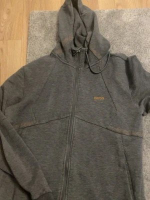 Grå hoodie från BOSS med dragkedja - Snygg grå hoodie från BOSS med dragkedja framtill och justerbar huva. Diskret BOSS-logga i guld på bröstet och praktiska fickor på sidorna. Tillverkad i mjukt material som känns skönt mot huden. Perfekt för en avslappnad och stilren look.