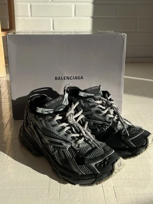 Balenciaga runner sneakers svart/grå - Säljer ett par Balenciaga runner sneakers i svart och grått med chunky sula och meshdetaljer. Skorna har flera lager av material, reflekterande detaljer och kraftiga snören. Märkeslogga på plös och häl. Perfekta för dig som gillar statement sneakers.