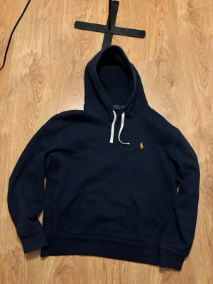 Mörkblå hoodie från Polo Ralph Lauren - Mörkblå hoodie från Polo Ralph Lauren med klassisk huva och vit. Tillverkad i mjukt bomullsmaterial och har en magficka framtill. Perfekt för en avslappnad och stilren look.